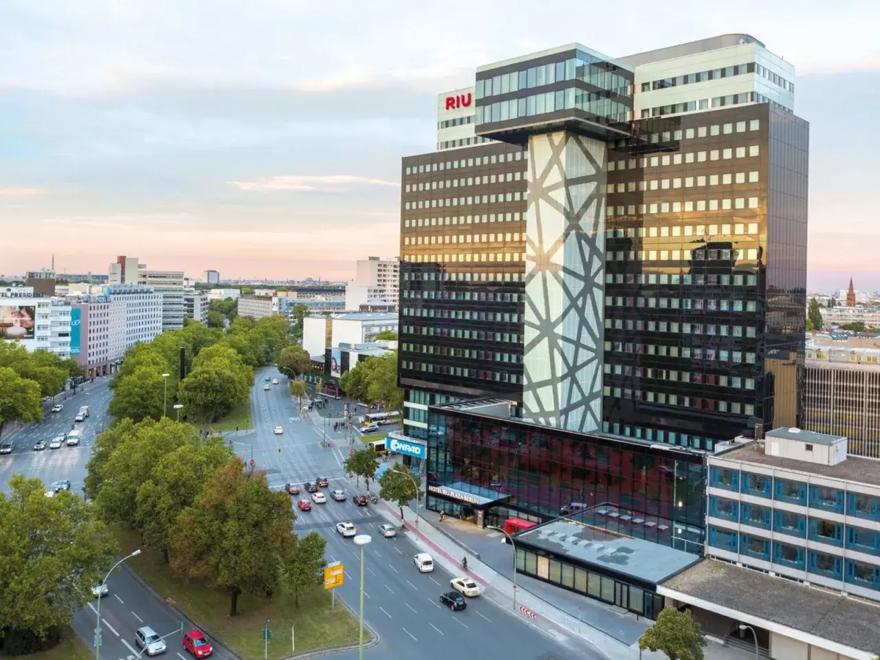 Das Hotel Riu Plaza Berlin, ein markantes Gebäude mit Glasfassade und geometrischem Muster, ragt über eine belebte Straße mit Bäumen und Verkehr.