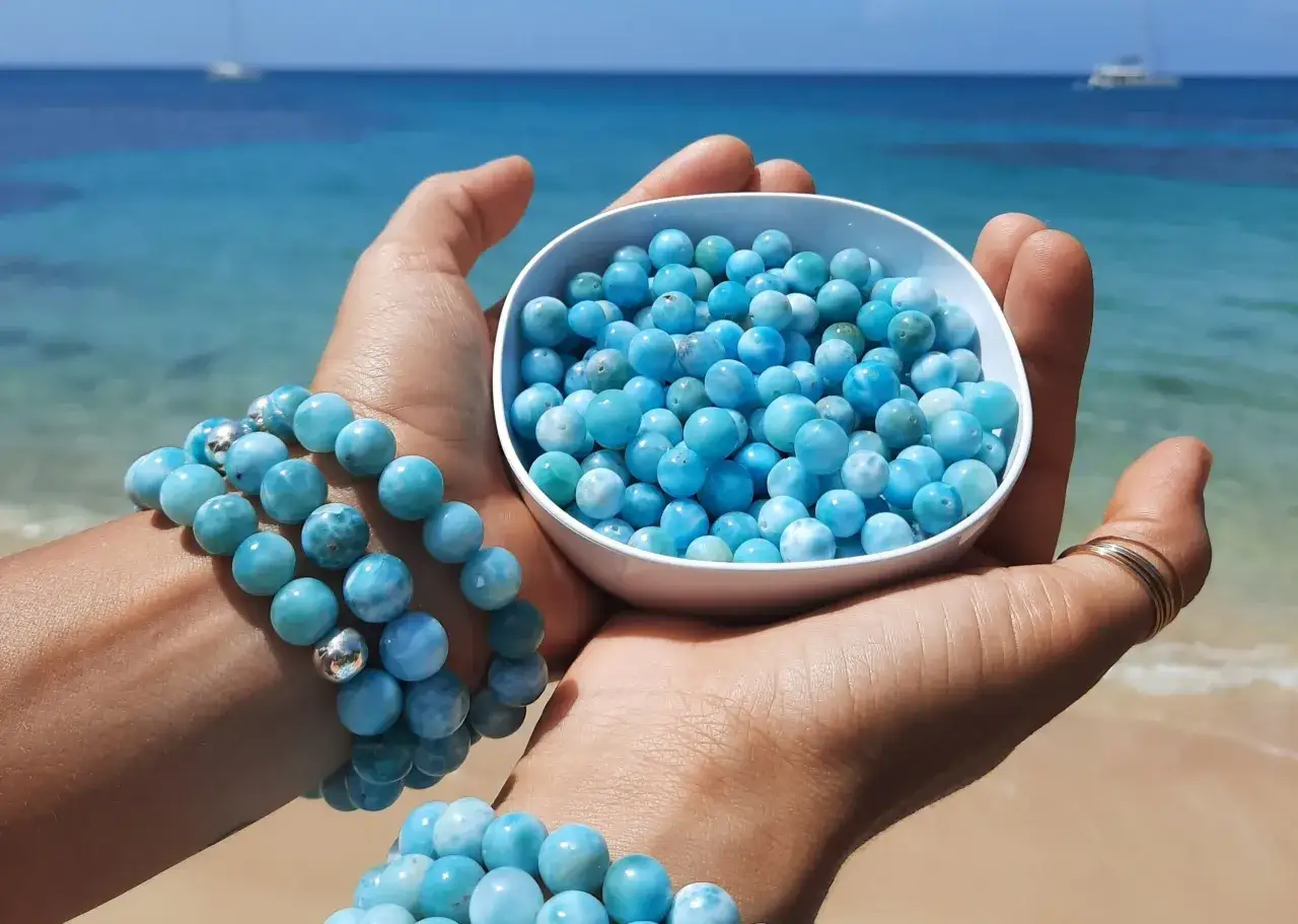 Mani che tengono una ciotola piena di perle di larimar, con braccialetti di larimar al polso. Lo sfondo è un mare azzurro e una spiaggia sabbiosa.