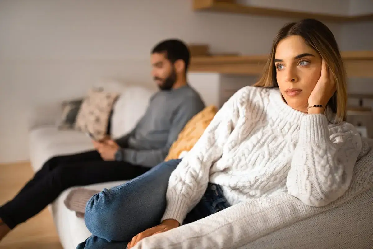 Un couple s'ennuie dans son couple, chacun absorbé par son téléphone sur le canapé, le regard perdu dans le vide.