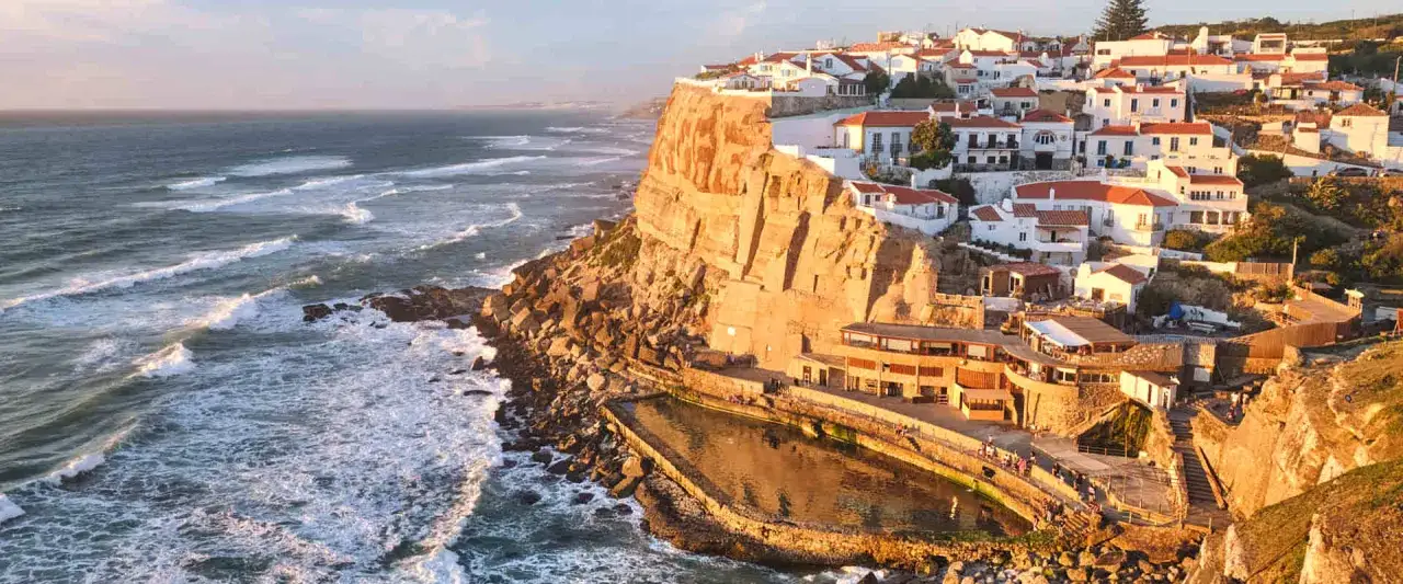 Coastal village with white houses and red roofs perched on a cliff overlooking the Atlantic Ocean. A natural swimming pool is carved into the rocks below. This scene captures the beauty of Portugal's Algarve coast.