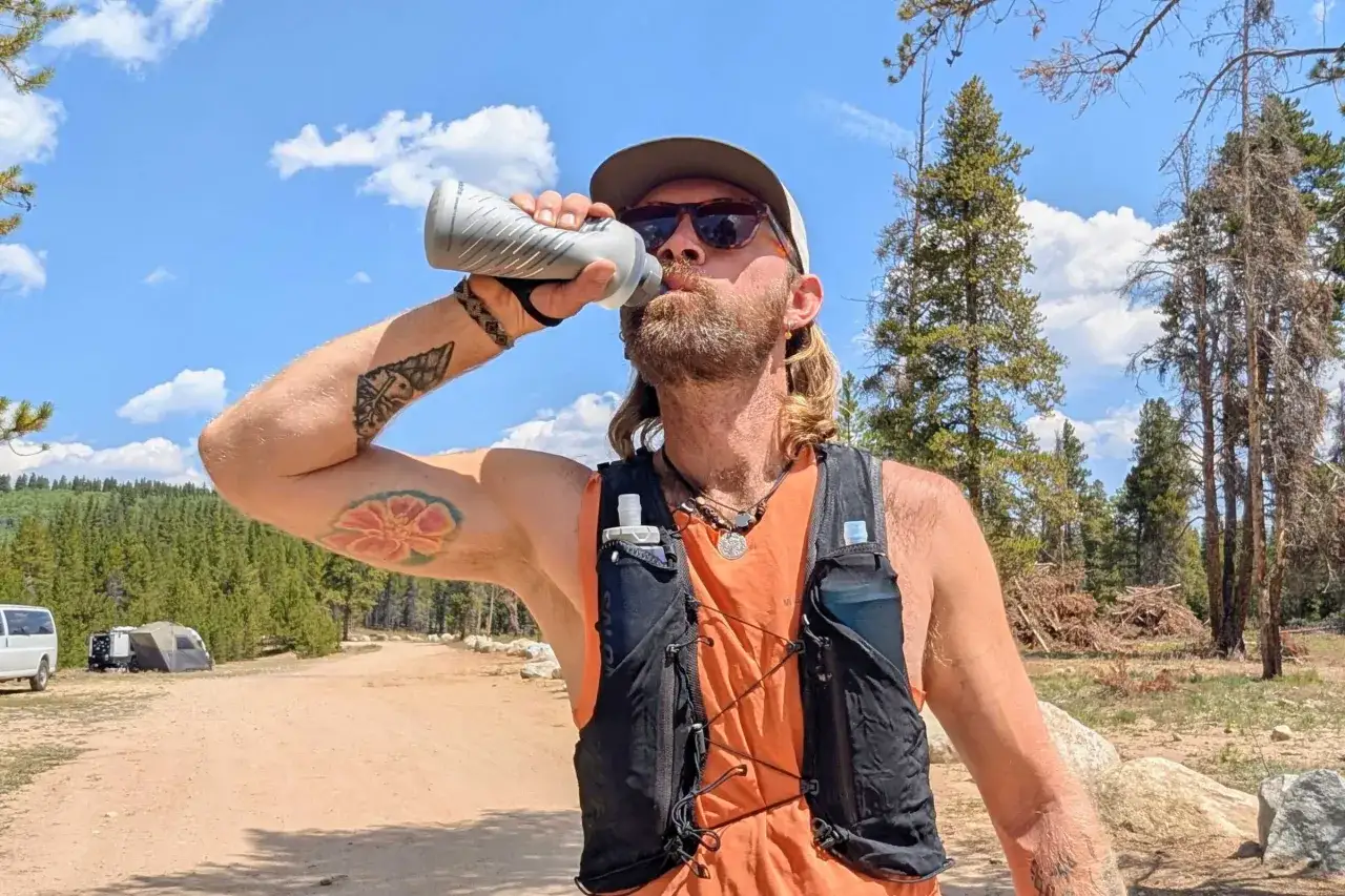 Un coureur boit dans sa gourde, essentiel pour l'hydratation sportif. Il porte un gilet de trail avec des poches pour l'eau, prêt pour sa course en pleine nature.