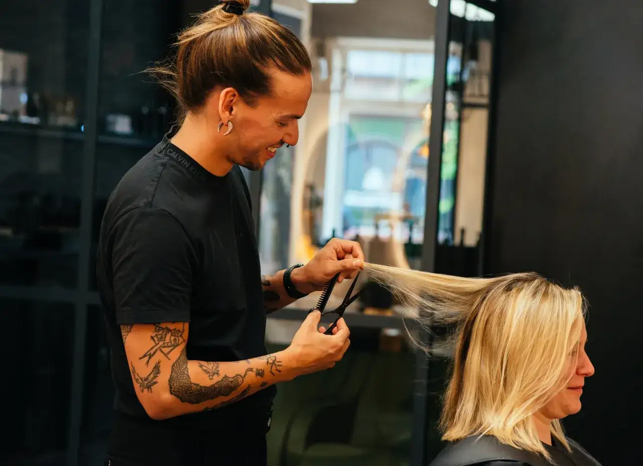 Friseur mit Tattoos schneidet einer Kundin die blonden Haare trocken.