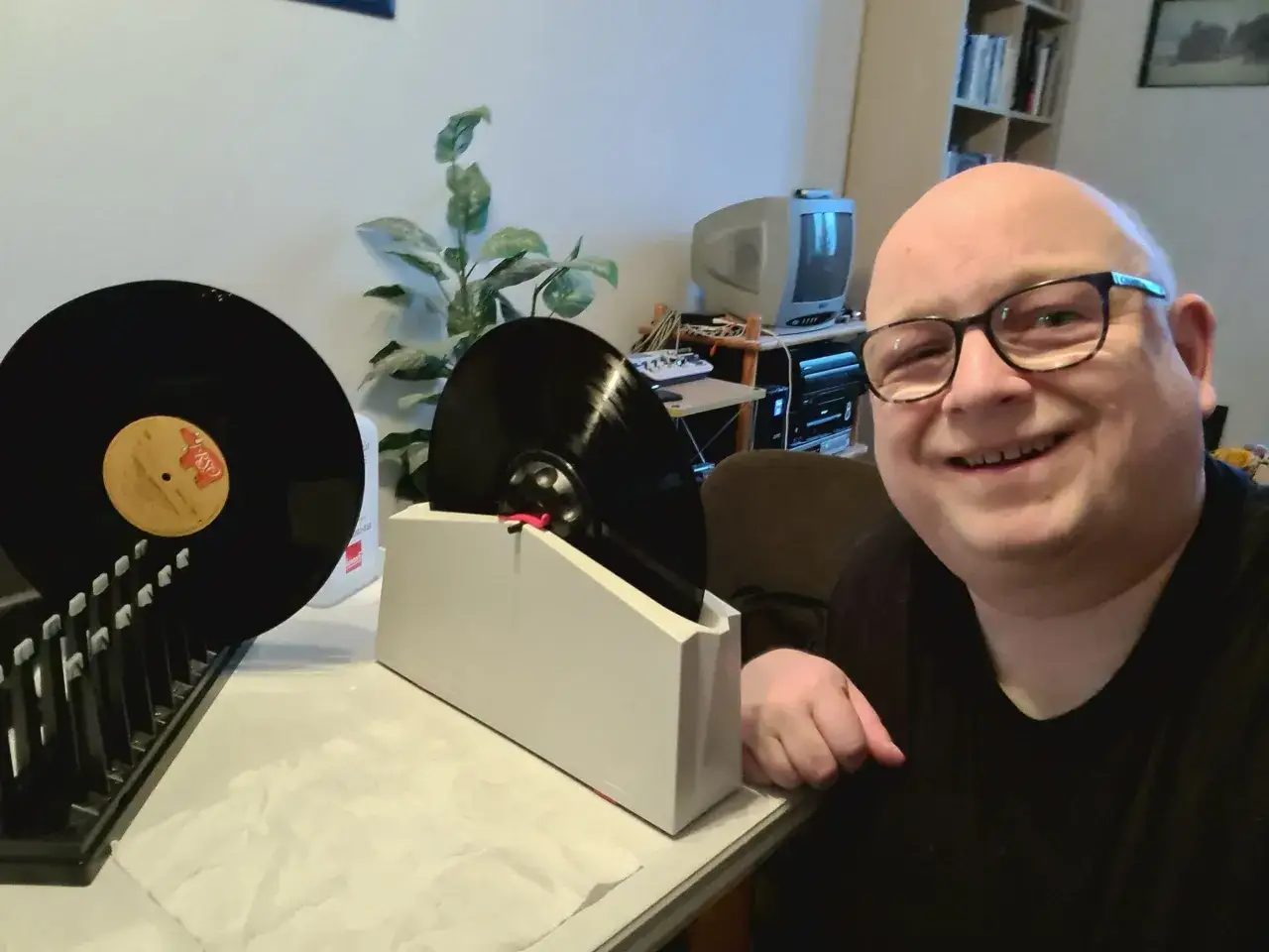 Mann lächelt, während er seine Schallplatten wäscht. Eine Schallplatte steht in einer Halterung, eine weitere liegt daneben.