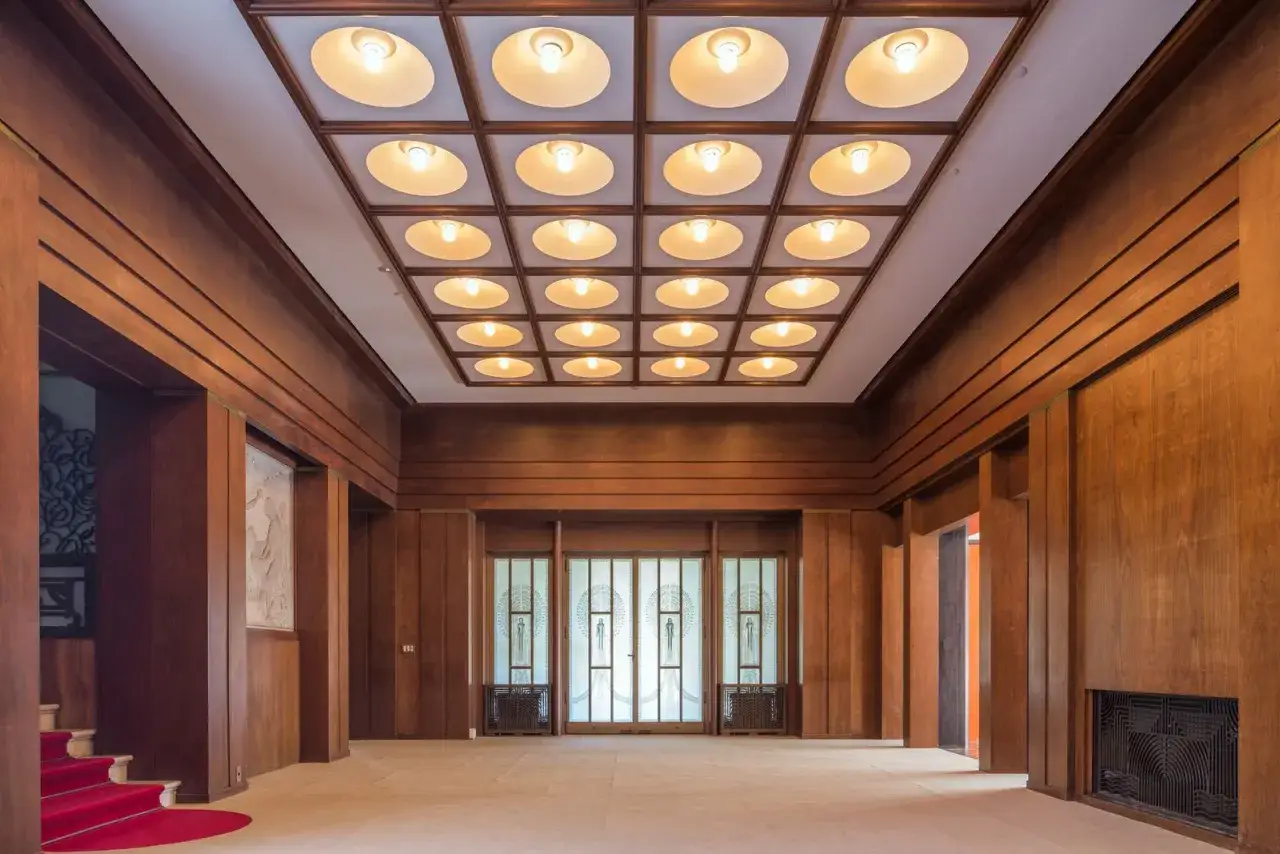 Interior of a grand hall with wood paneling, a red carpeted staircase, and a grid of illuminated ceiling lights, reminiscent of the elegant design found in some Japan art museums.