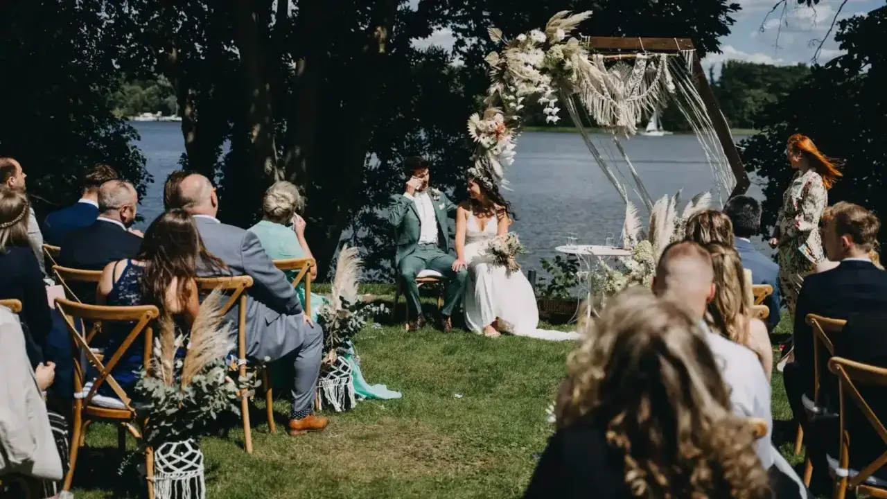 Hochzeit am See mit viel greenery. Brautpaar sitzt unter einem Bogen, Gäste auf Stühlen.