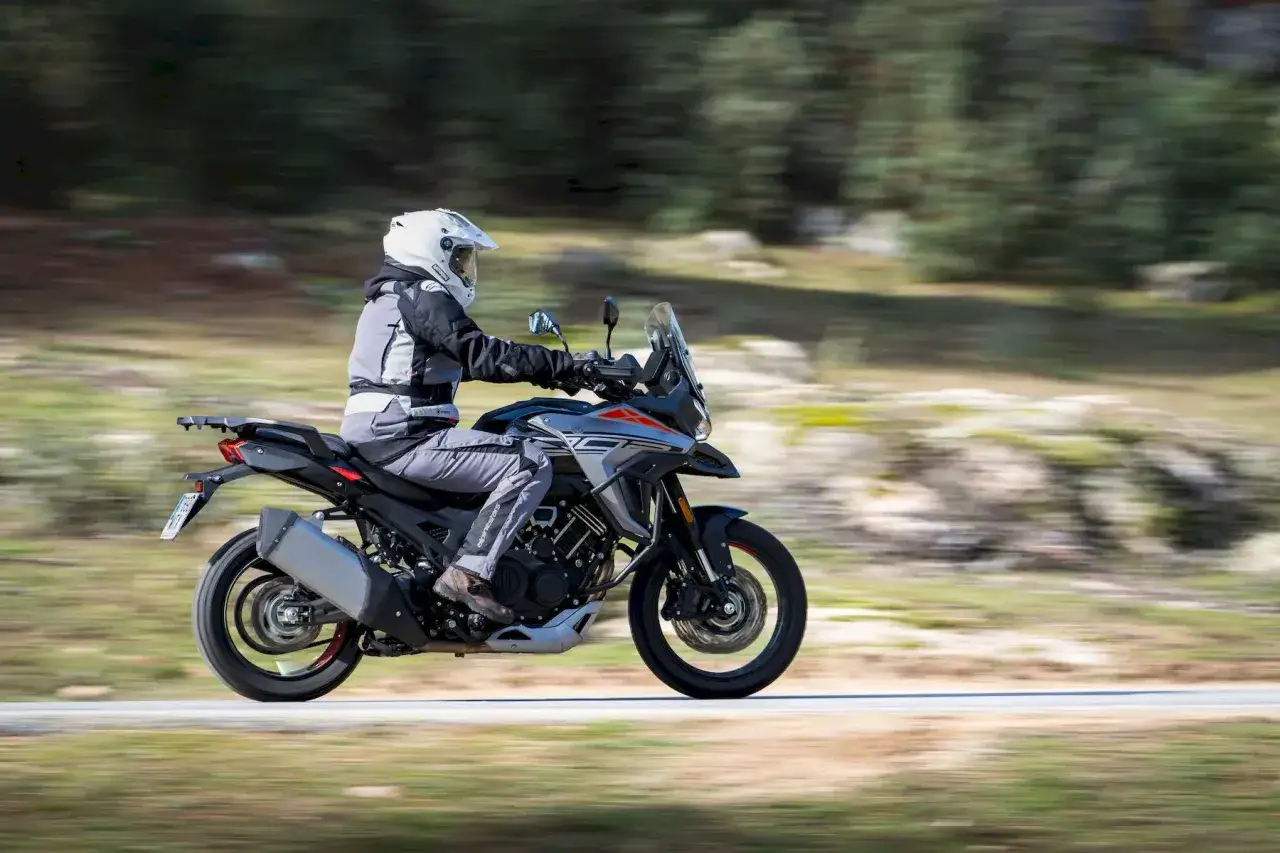 Motociclista con casco blanco y equipo de aventura conduce una moto china trail por un camino rural con árboles borrosos al fondo.