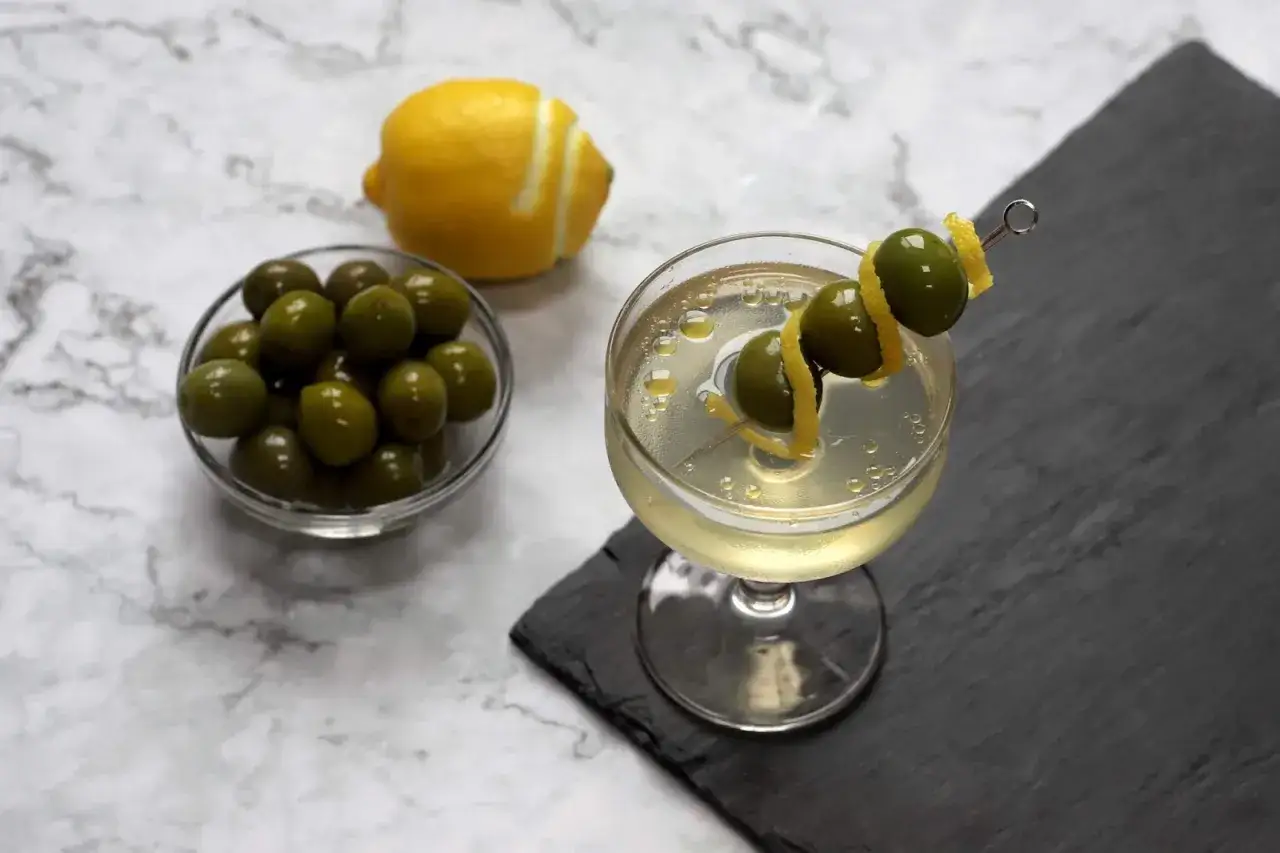 Un verre de martini avec trois olives sur une brochette et un zeste de citron. À côté, un bol d'olives et un citron.