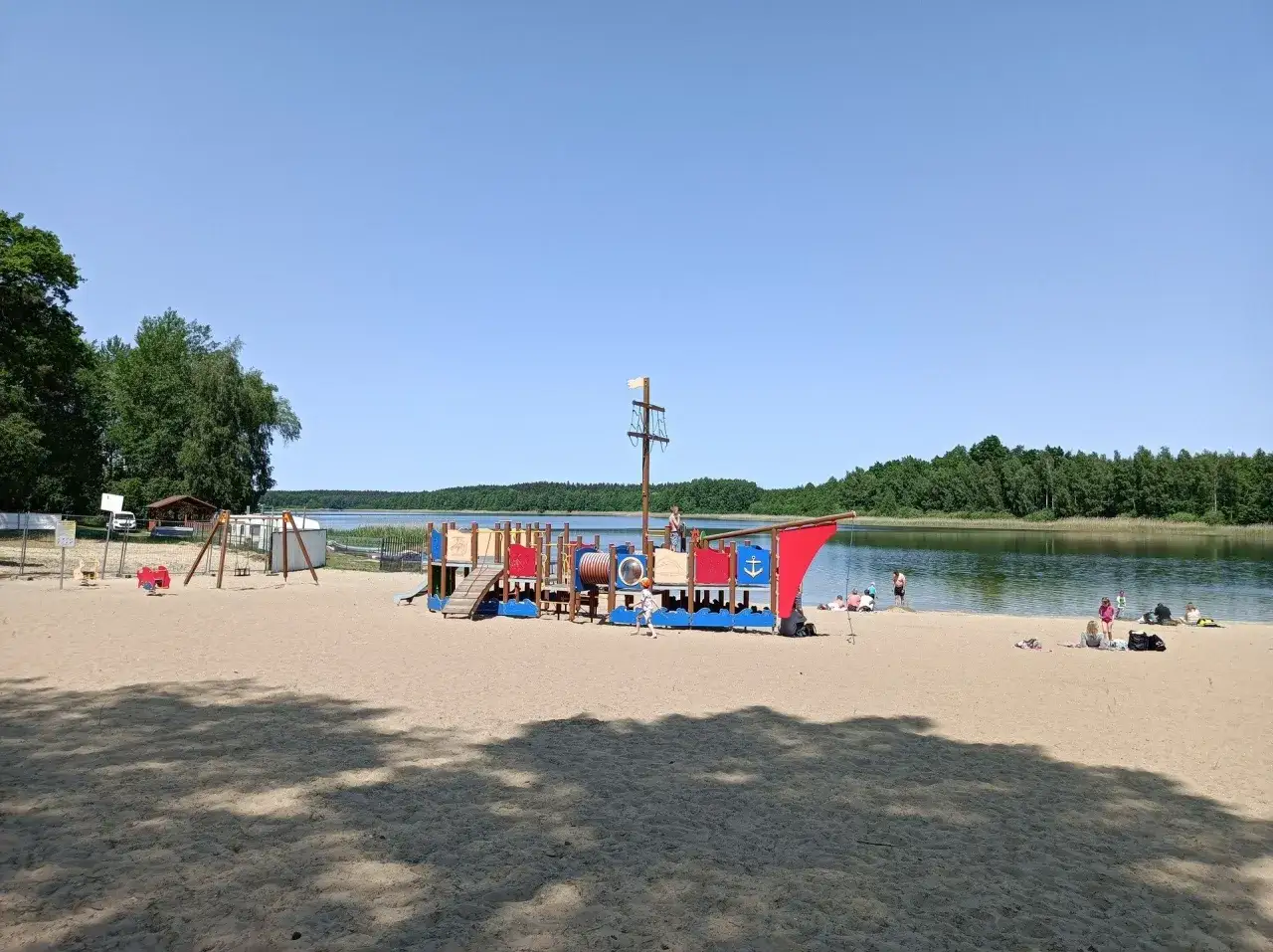 Plaża nad jeziorem Kamionki z placem zabaw w kształcie statku pirackiego. Dzieci bawią się na piasku, a w tle widać spokojną taflę wody i zielone drzewa.