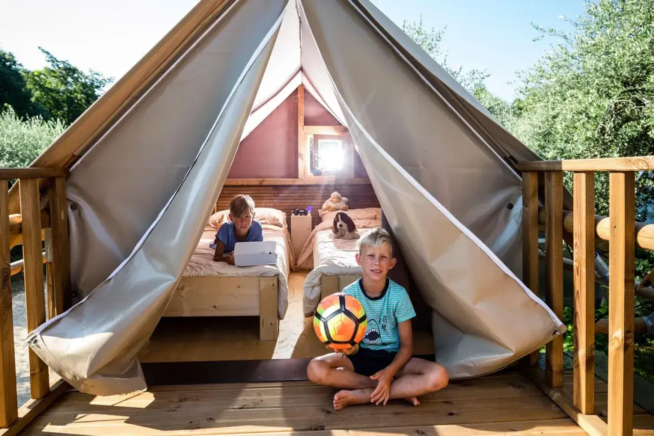 Bambini felici in un accogliente glamping per famiglie. Uno gioca con un pallone, l'altro usa un tablet a letto.