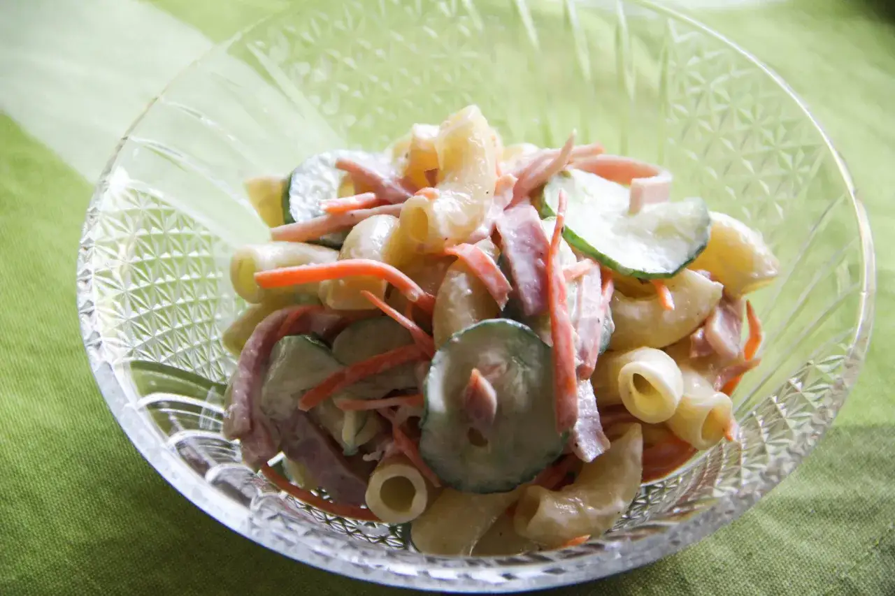 A close-up of a glass bowl filled with creamy Japanese macaroni salad, featuring elbow pasta, shredded carrots, cucumber slices, and ham.