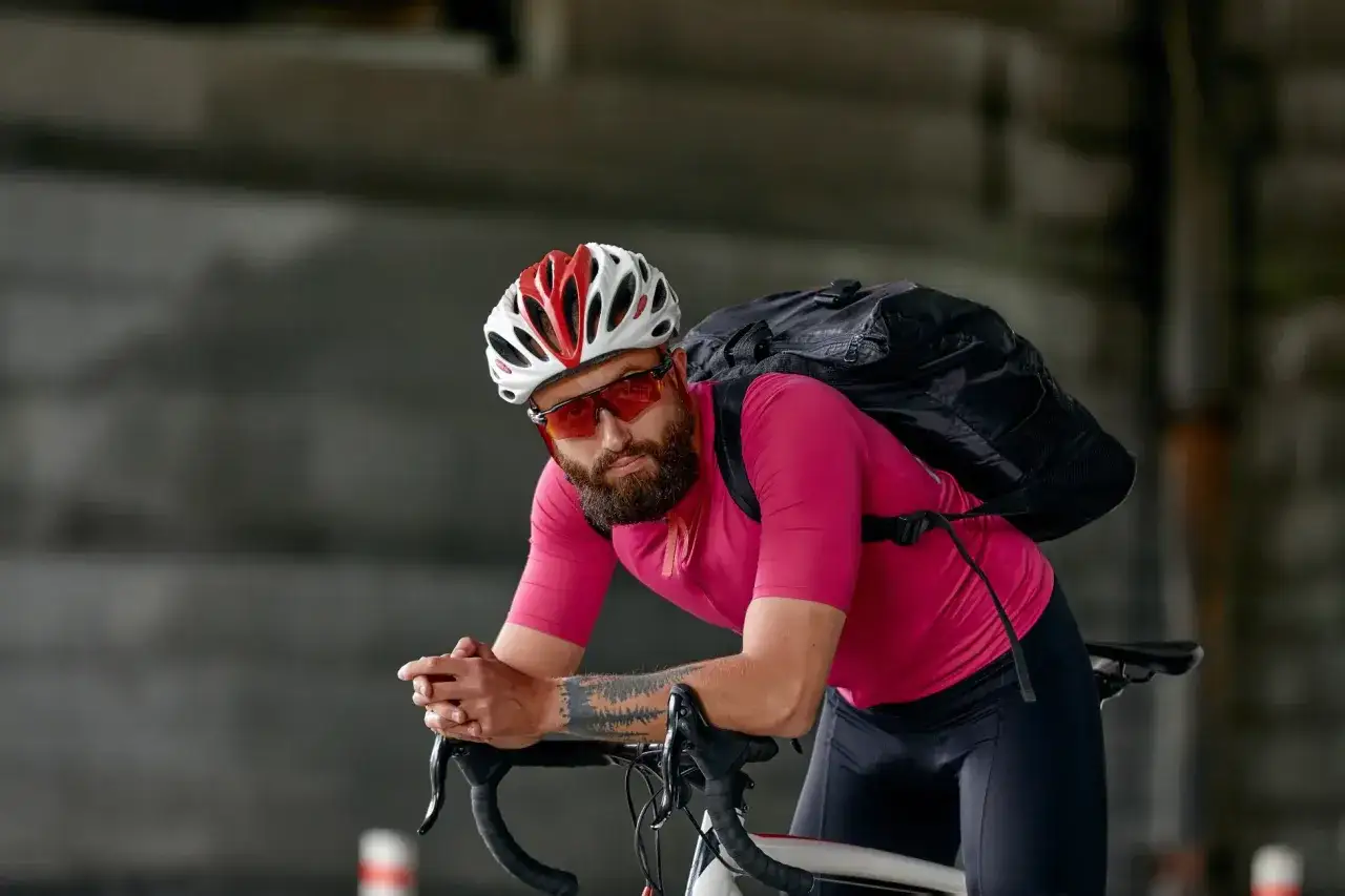 Mężczyzna w kasku, okularach i różowej koszulce, z plecakiem na plecach, opiera się o kierownicę roweru. Kompletne wyposażenie rowerzysty.