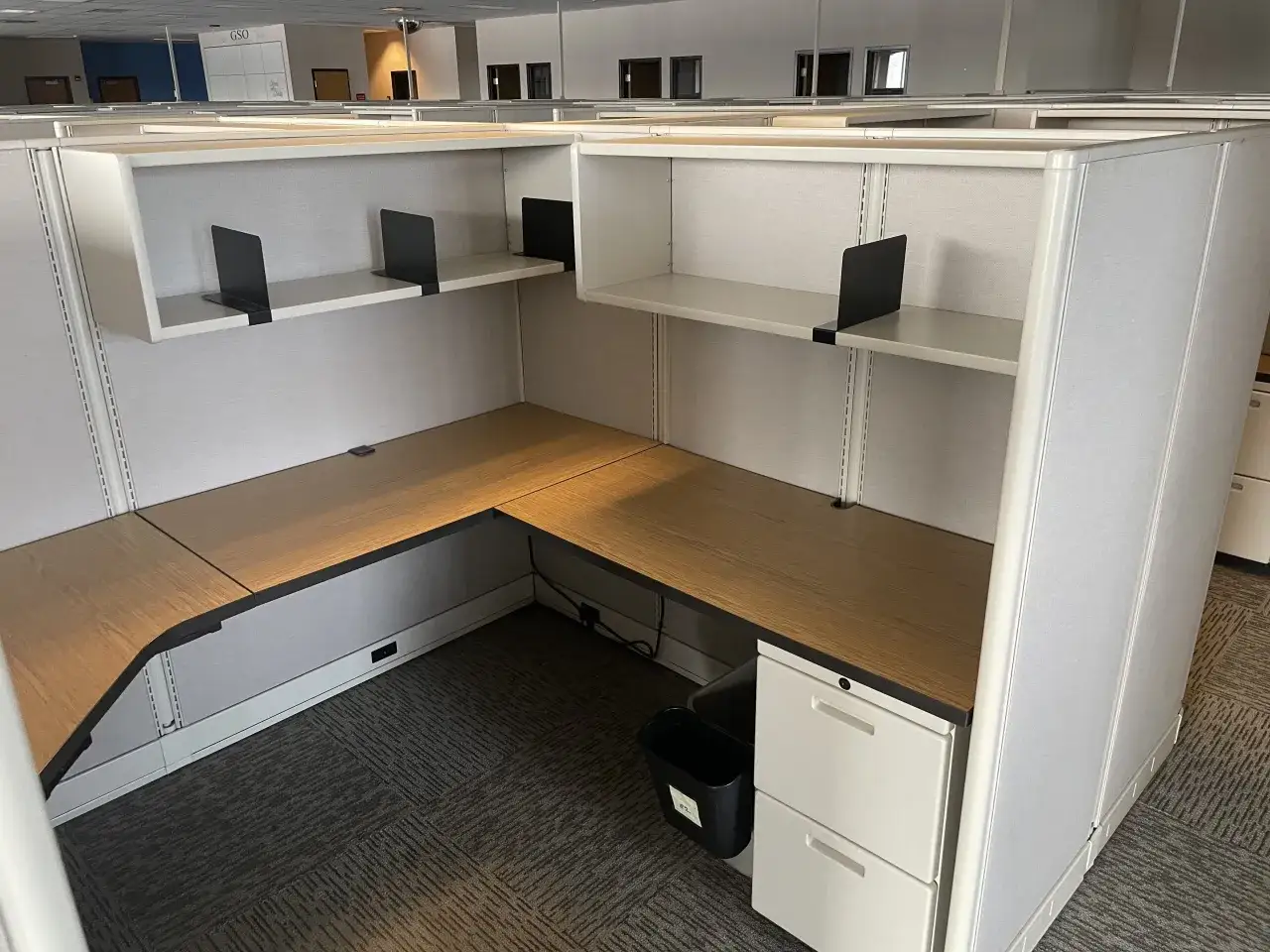 Leerer Bürokubus mit L-förmigem Schreibtisch, ideal für Feng Shui. Helle Wände, Holzoberfläche und weiße Schubladen.