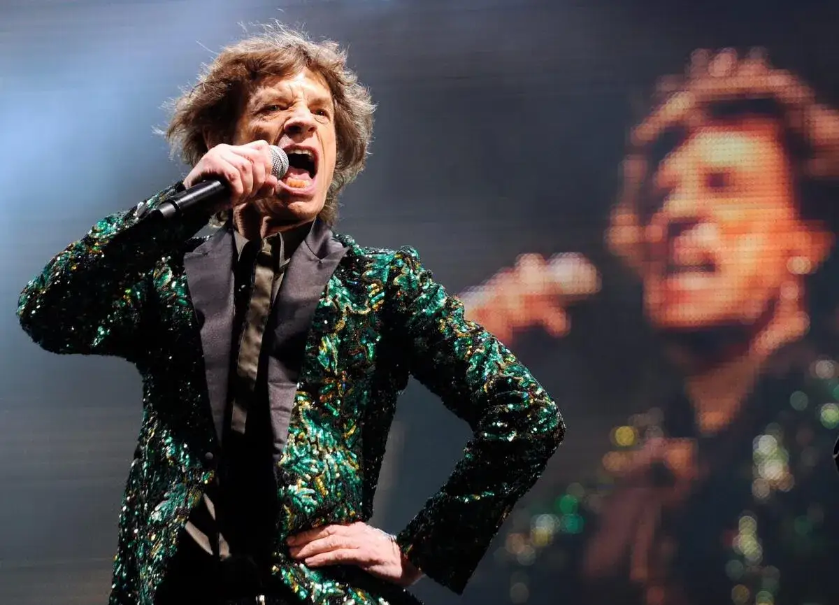 Mick Jagger, w błyszczącej marynarce, śpiewa na scenie. W tle widać jego powiększony obraz. To ikona zespołu The Rolling Stones.