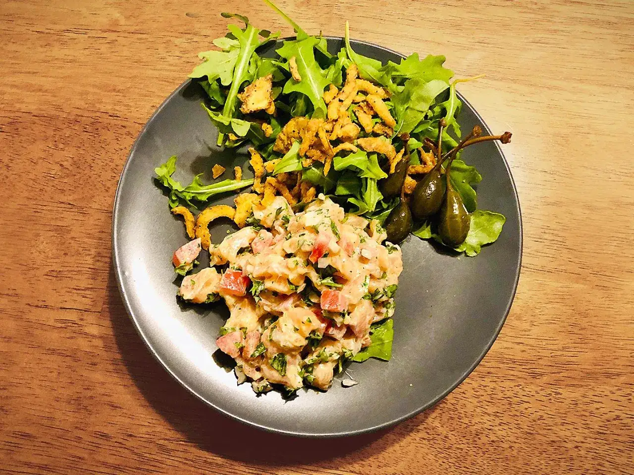 D&eacute;licieux tartare de saumon frais, servi avec une salade croquante, des c&acirc;pres et des oignons frits sur une assiette noire.
