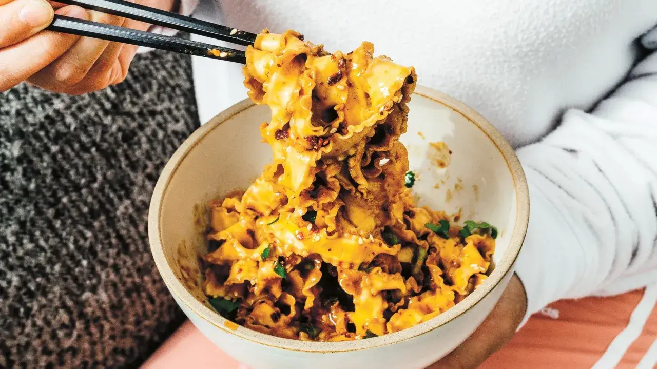 Chopsticks unoszą faliste kluski oblane pikantnym, pomarańczowym sosem do makaronu po chińsku, posypane sezamem i szczypiorkiem.