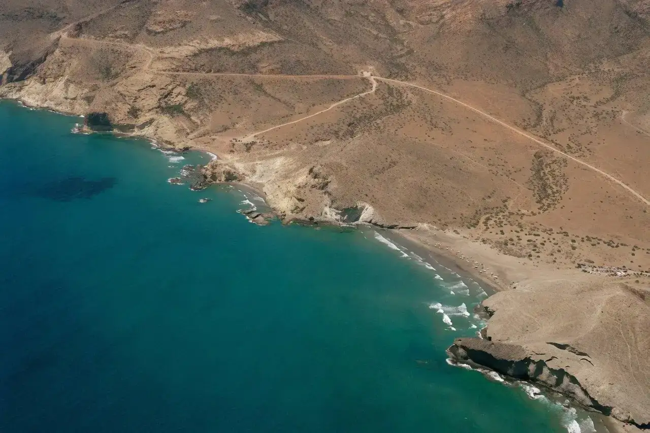 Vista aérea de la cala de la media luna, con aguas turquesas y acantilados áridos. Un camino serpentea por la colina.