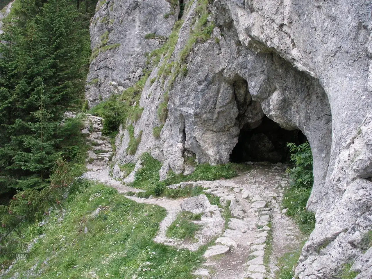Kamienista ścieżka prowadzi do wejścia do jaskiń w górach. Wokół bujna zieleń i skaliste zbocza.