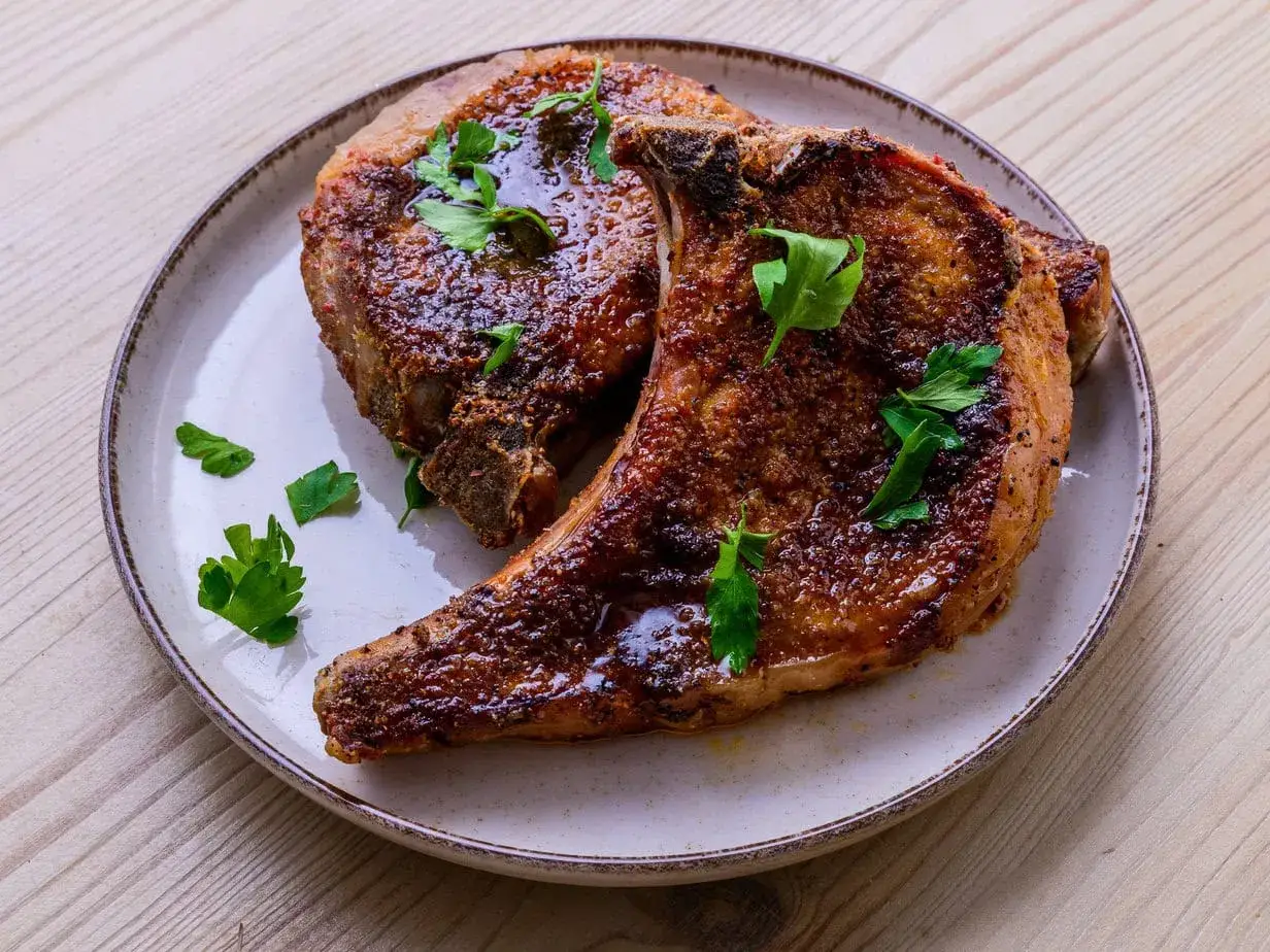 Deux belles c&ocirc;tes de porcelet grill&eacute;es, napp&eacute;es d'une sauce brillante et garnies de persil frais, sur une assiette.
