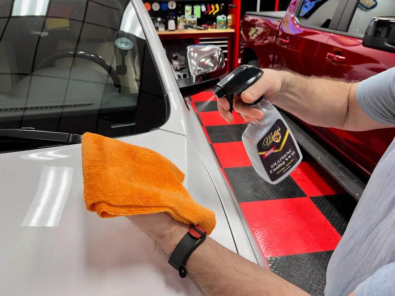En hand sprutar spray wax på en vit bil. En orange mikrofiberduk ligger redo för att polera.