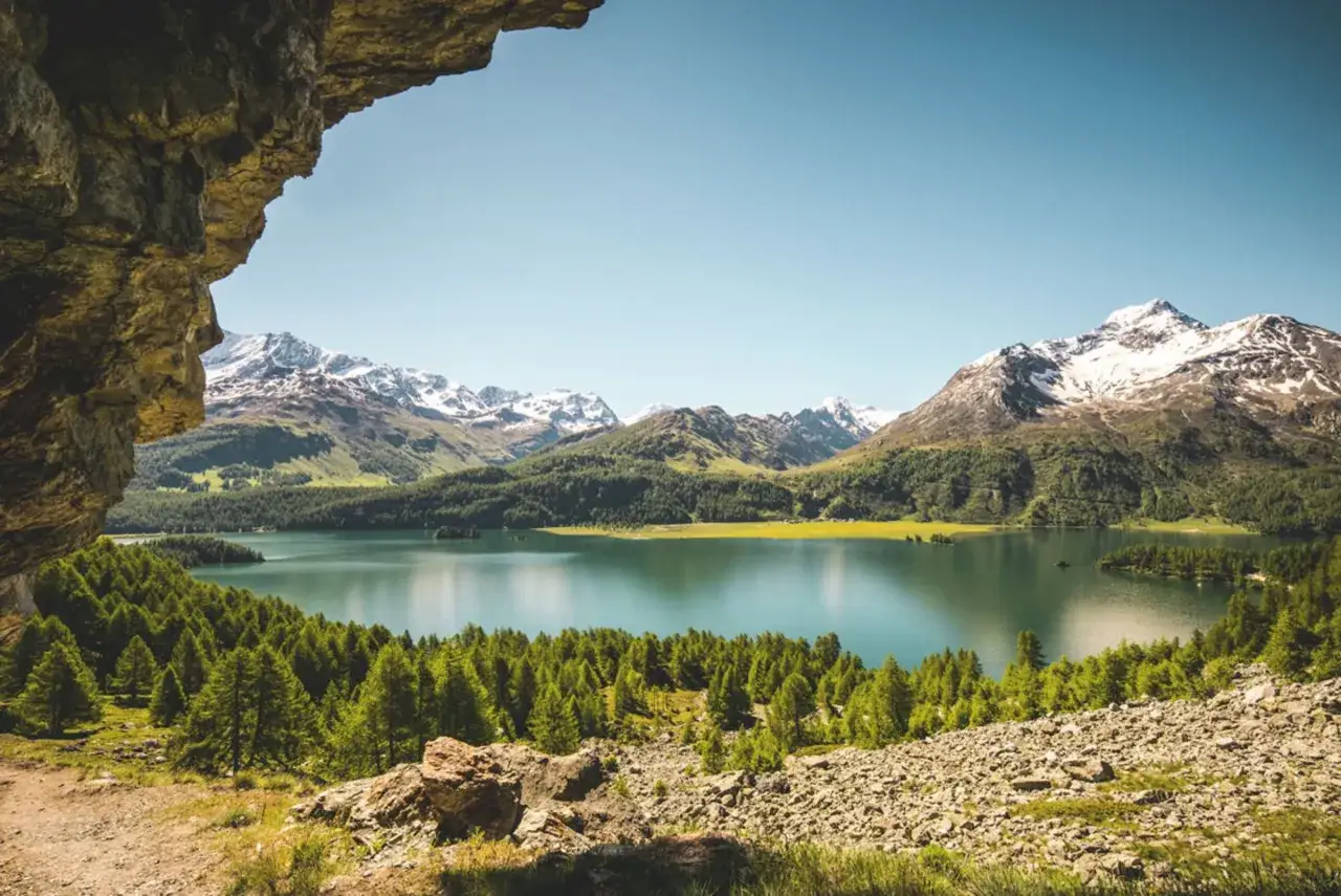 Felsiger Aussichtspunkt mit Blick auf einen türkisfarbenen See, umgeben von grünen Wäldern und schneebedeckten Bergen. Ein malerischer Ausblick, der an die Grenze zwischen Schweiz und Italien erinnert.