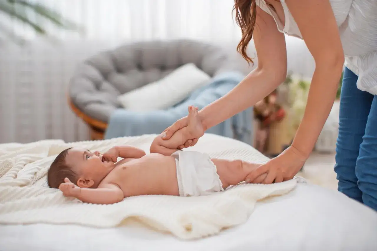 Mama delikatnie podnosi nóżkę noworodka, sprawdzając, co ile zmieniać pieluszkę. Maluch leży na miękkim kocyku.