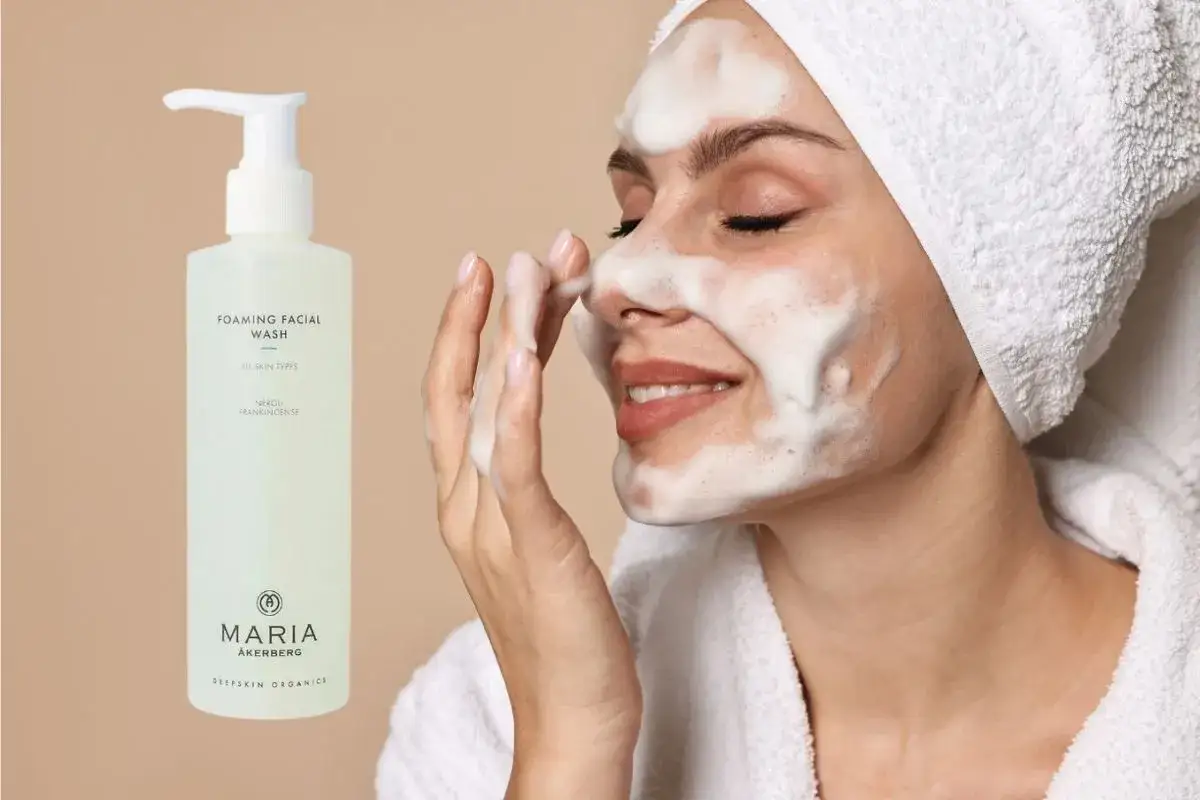 En kvinna med skum i ansiktet använder Maria Åkerberg foaming facial wash. En flaska av produkten syns till vänster.
