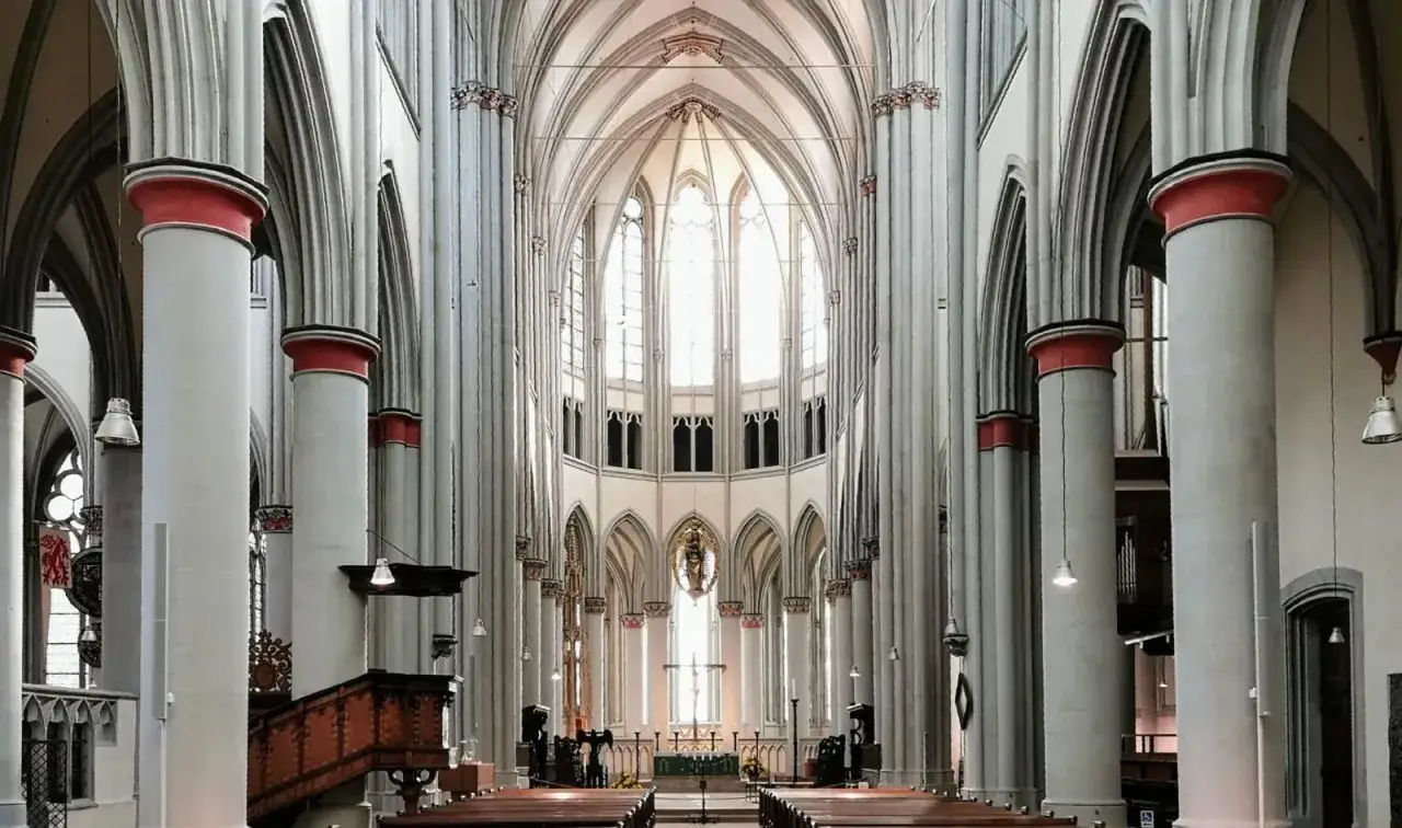 Hohe S&auml;ulen mit roten Akzenten und Spitzb&ouml;gen im Altenberger Dom. Das Licht f&auml;llt durch die gro&szlig;en Fenster im Hintergrund.