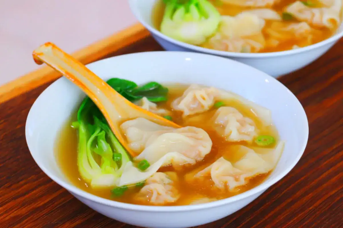 Aksamitna zupa wonton z zielonymi warzywami i pomarańczową łyżką, podana w białej misce na drewnianym tle.