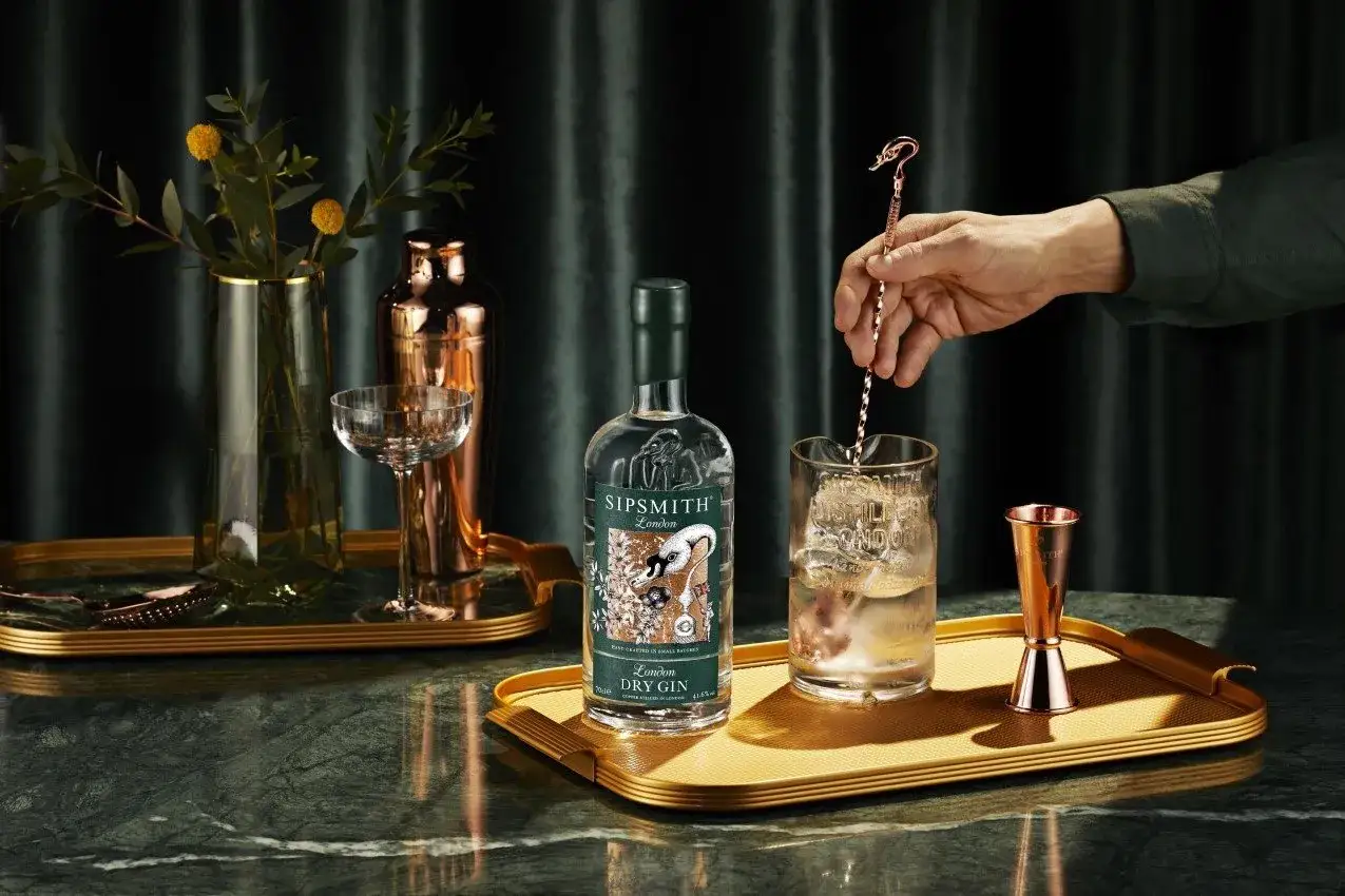 En hand rör om i ett glas med is och en drink med gin. En flaska Sipsmith London Dry Gin står bredvid.