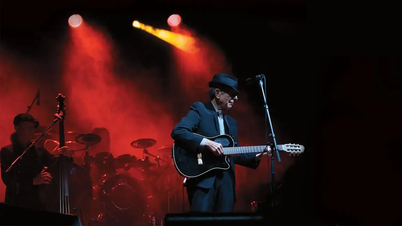 Leonard Cohen spielt Gitarre auf der B&uuml;hne, umgeben von rotem B&uuml;hnenlicht. Ein Kontrabassist und Schlagzeuger sind im Hintergrund zu sehen.