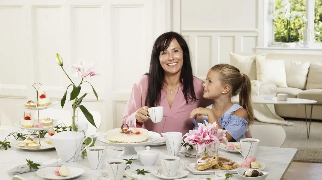 Uśmiechnięta mama i córka celebrują Dzień Mamy przy stole pełnym słodkości i kawy.