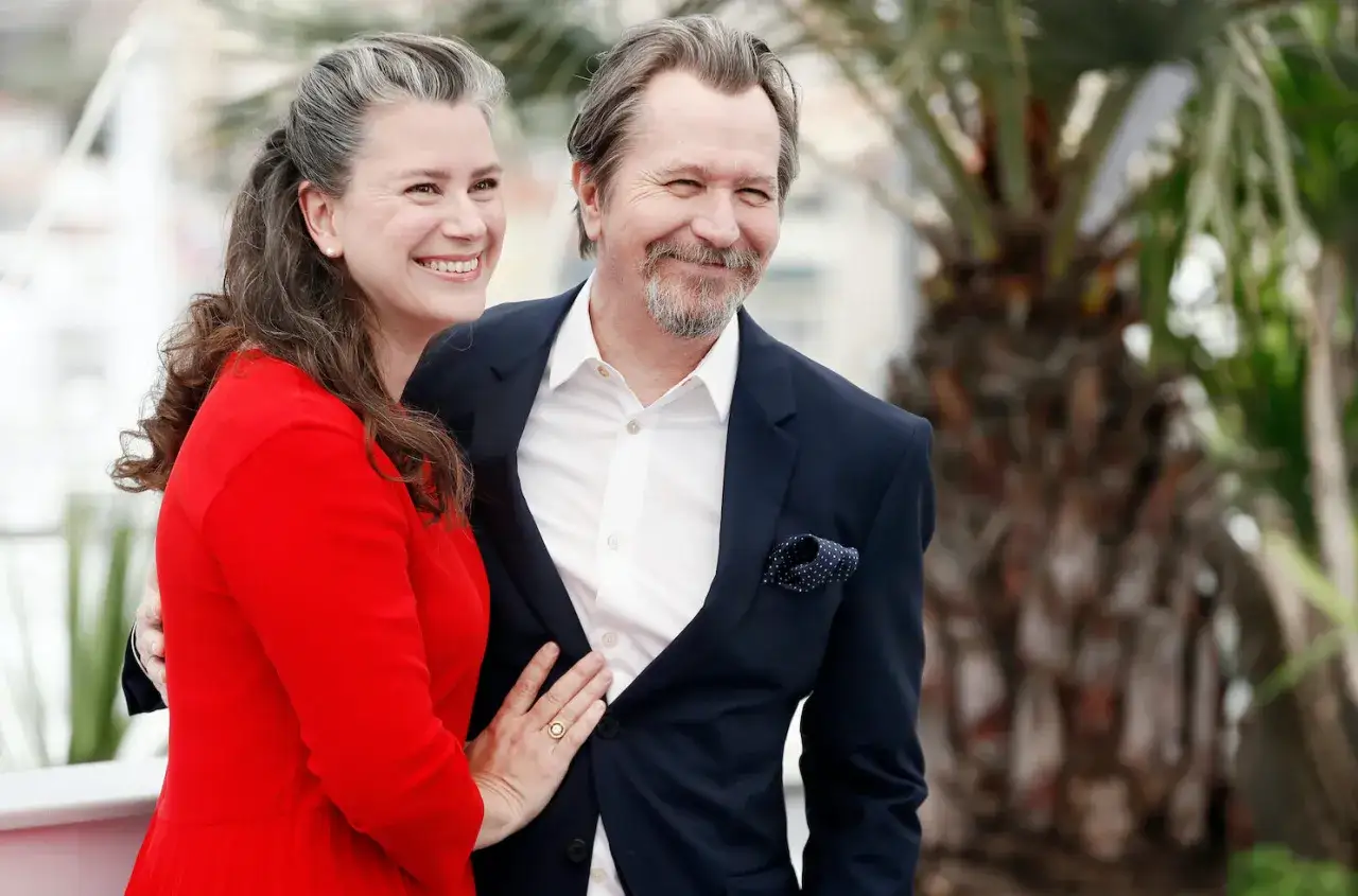 Gary Oldman z żoną, uśmiechnięci, pozują do zdjęcia. Ona w czerwonej sukience, on w granatowym garniturze.