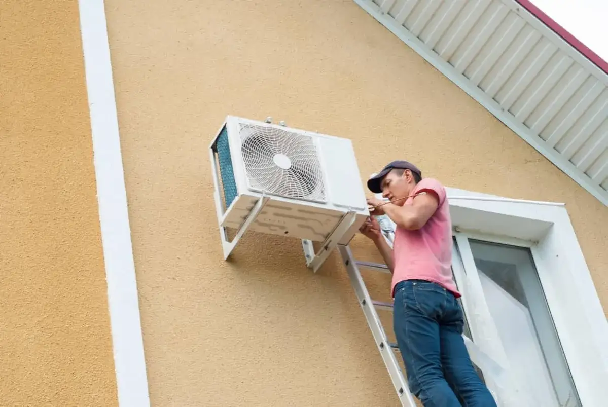 Técnico en escalera, trabajando en cómo instalar aire acondicionado en la pared exterior de una casa.