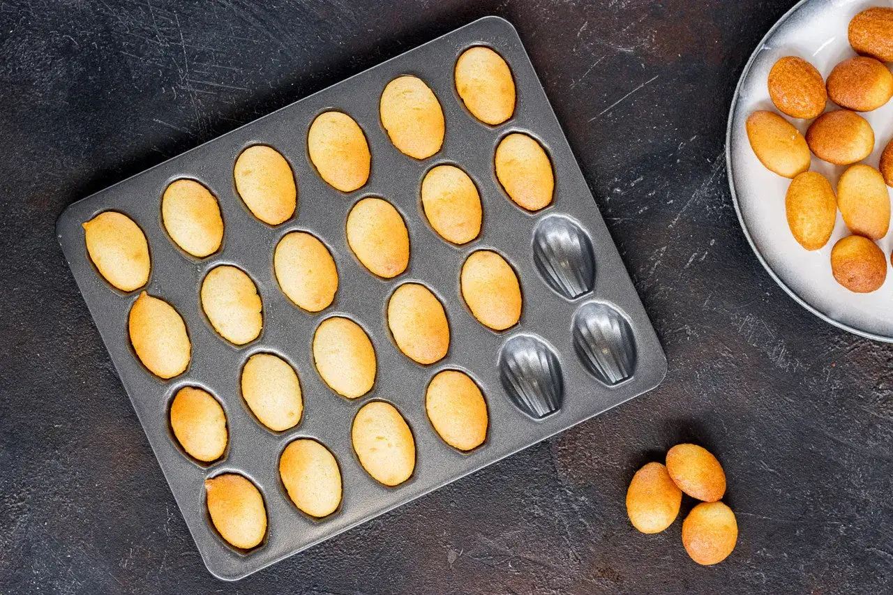 Frisch gebackene Keks-Madeleines in einer Backform, bereit für das magdalenas rezept. Ein paar liegen auf einem Teller.