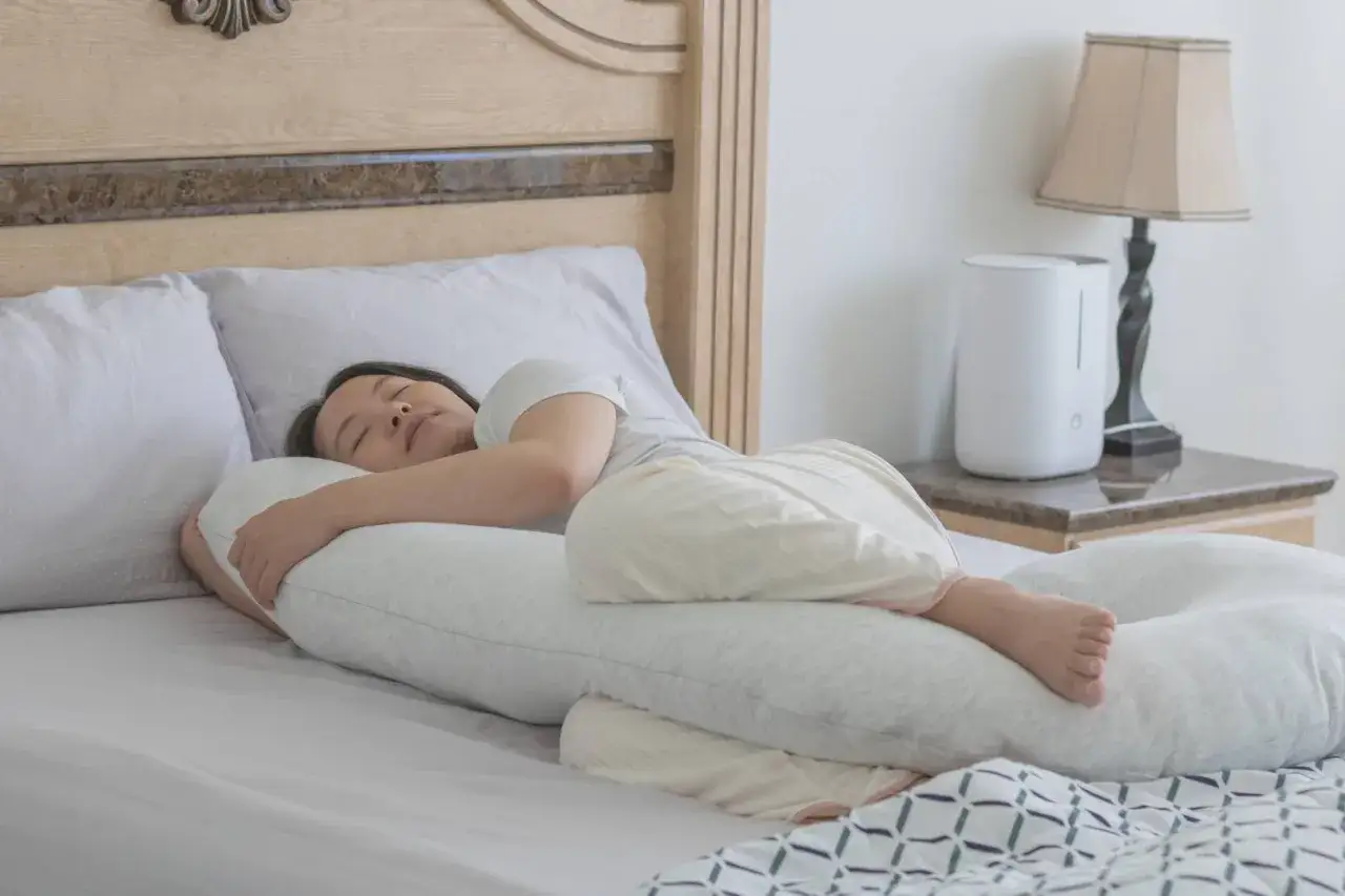A pregnant woman sleeps soundly, embracing a C-shaped pillow. This pillow is a great way to como aliviar el dolor de espalda en el embarazo, offering comfort and support.