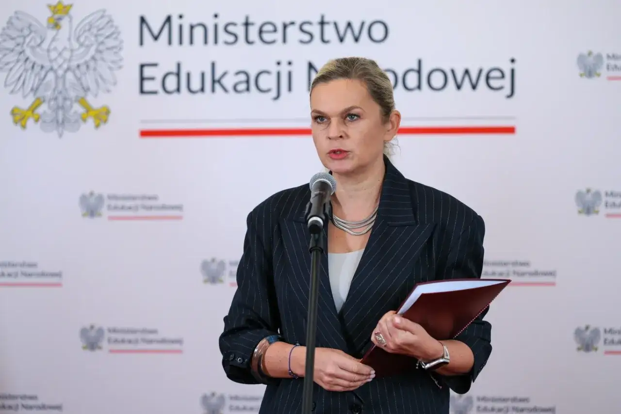 Minister edukacji przemawia przy mikrofonie, trzymając teczkę. W tle widnieje godło Polski i nazwa ministerstwa.