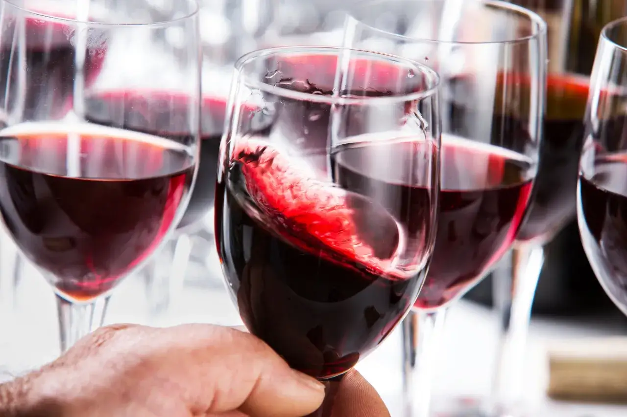 Ein Glas mit wirbelndem Grenache Rotwein wird von einer Hand gehalten. Mehrere Gl&auml;ser mit tiefrotem Wein sind im Hintergrund zu sehen.