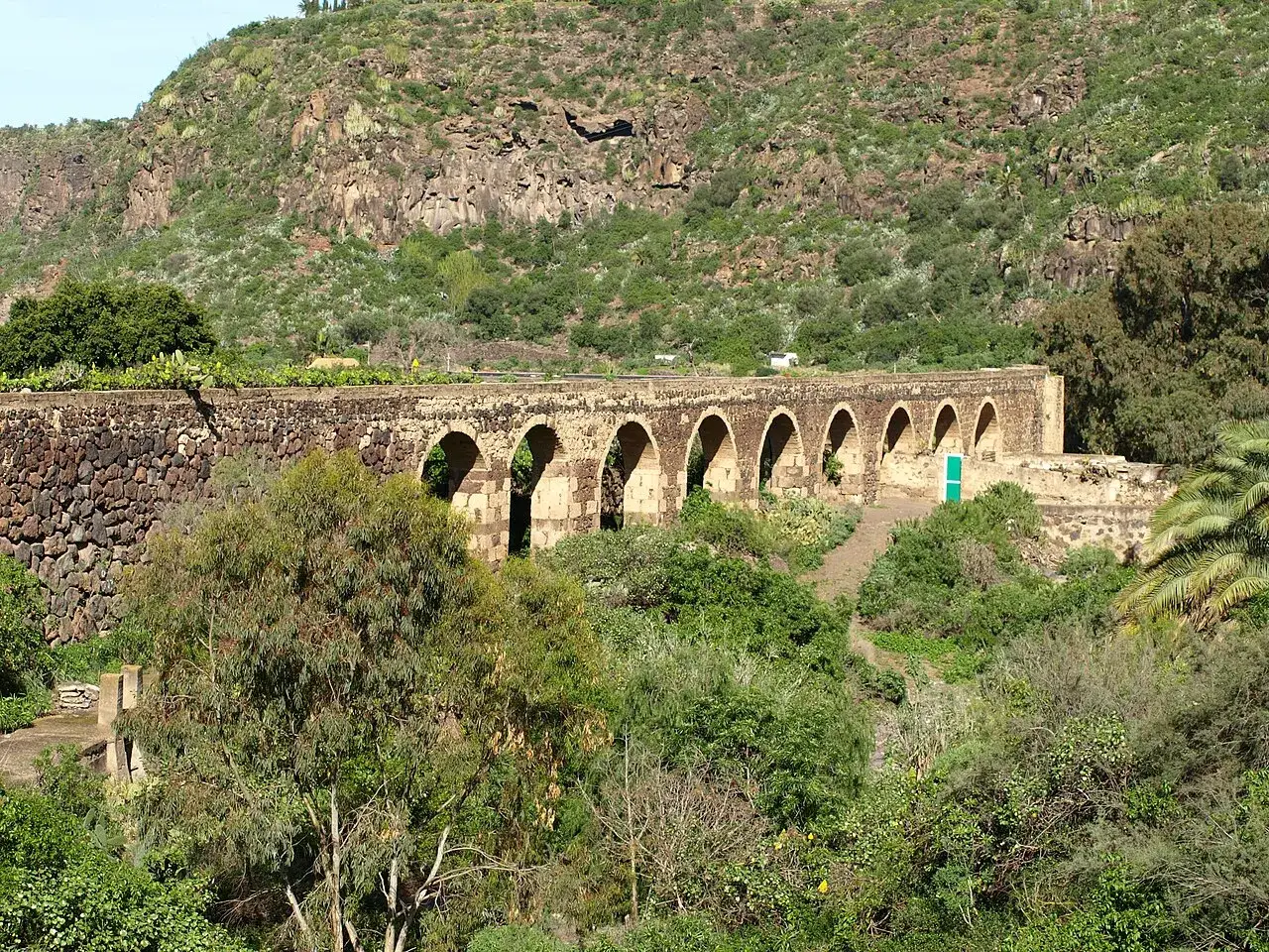 Acueducto de piedra con arcos en el barranco de Guiniguada, rodeado de vegetaci&oacute;n exuberante y una ladera rocosa al fondo.