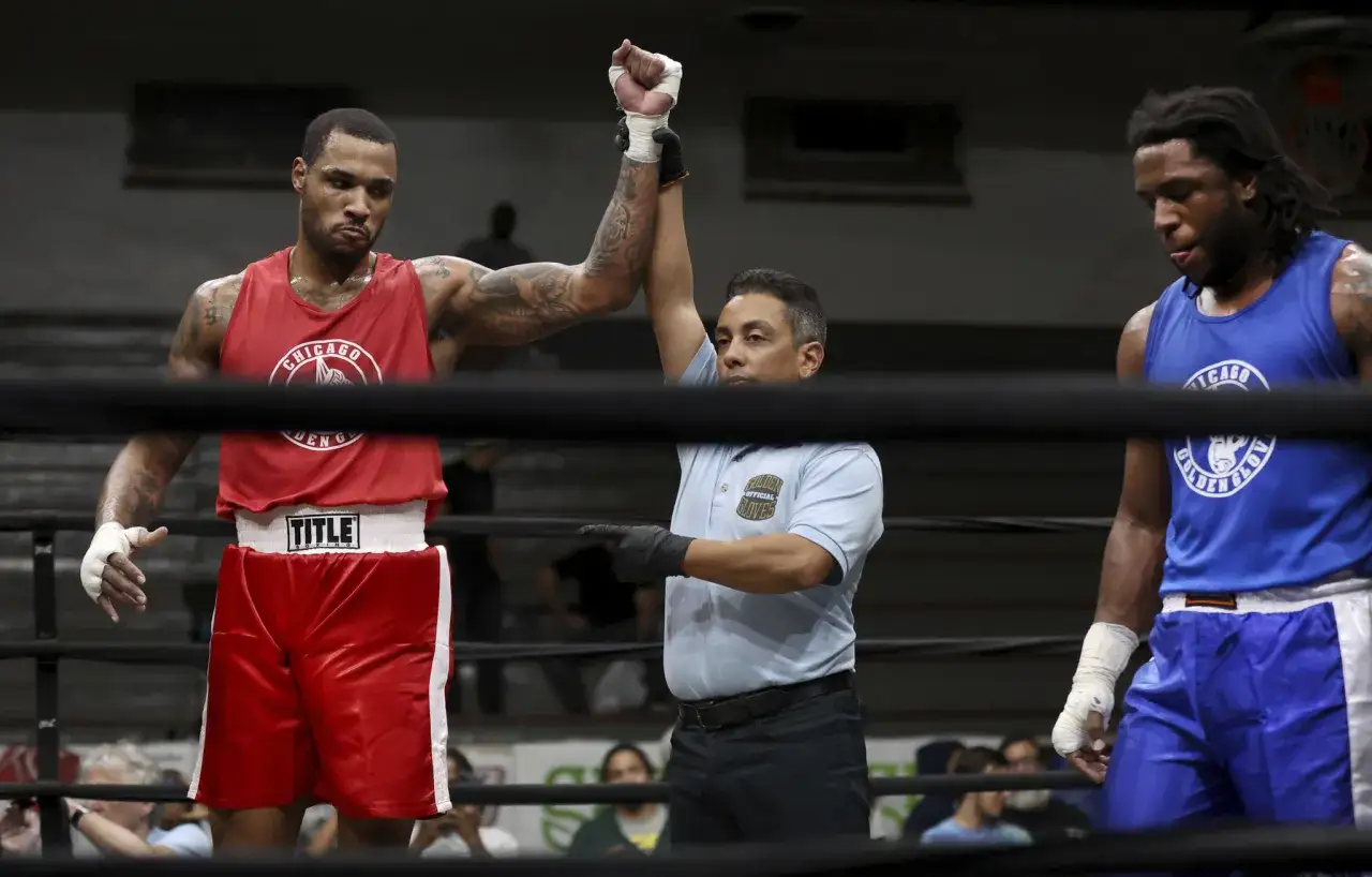 A referee raises the arm of a boxer in red shorts, signaling a win. The defeated boxer in blue shorts looks down. This is a super heavyweight weight boxing match.