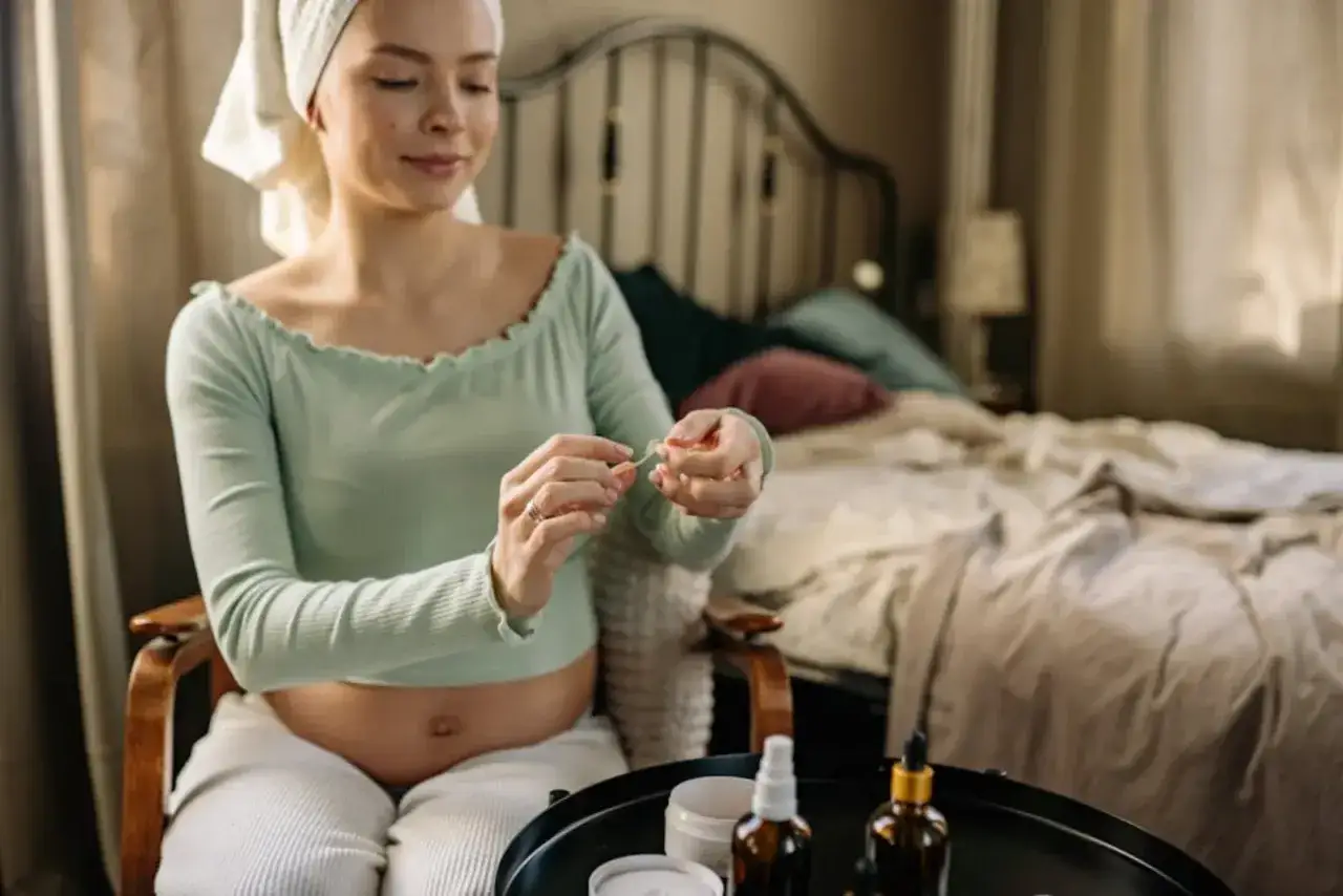 Gravid kvinna med handduk på huvudet tar hand om sin hud. Hon sitter i en stol med en säng i bakgrunden.