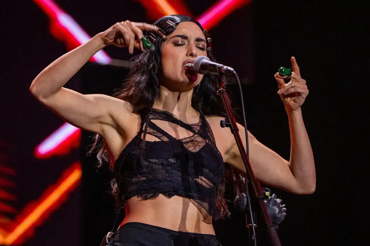 Une chanteuse aux cheveux noirs, portant des pinces à cheveux, chante dans un microphone. Son style de musique est intense.