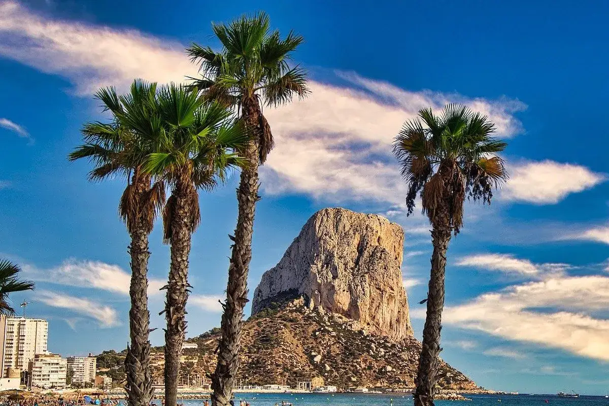 Palmeras en primer plano con el imponente Peñón de Ifach al fondo, destacando su altura y majestuosidad.