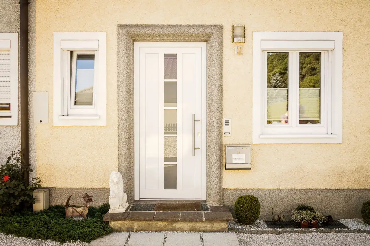 La entrada principal de una casa con una puerta blanca y ventanas a los lados. Los colores de puertas y ventanas contrastan con la fachada amarilla.