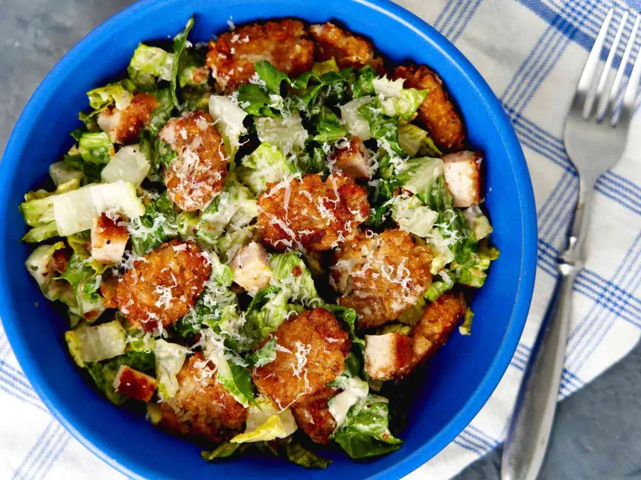 En bl&aring; sk&aring;l med caesarsallad, kycklingbitar och krispiga tater tots, toppad med riven parmesan.