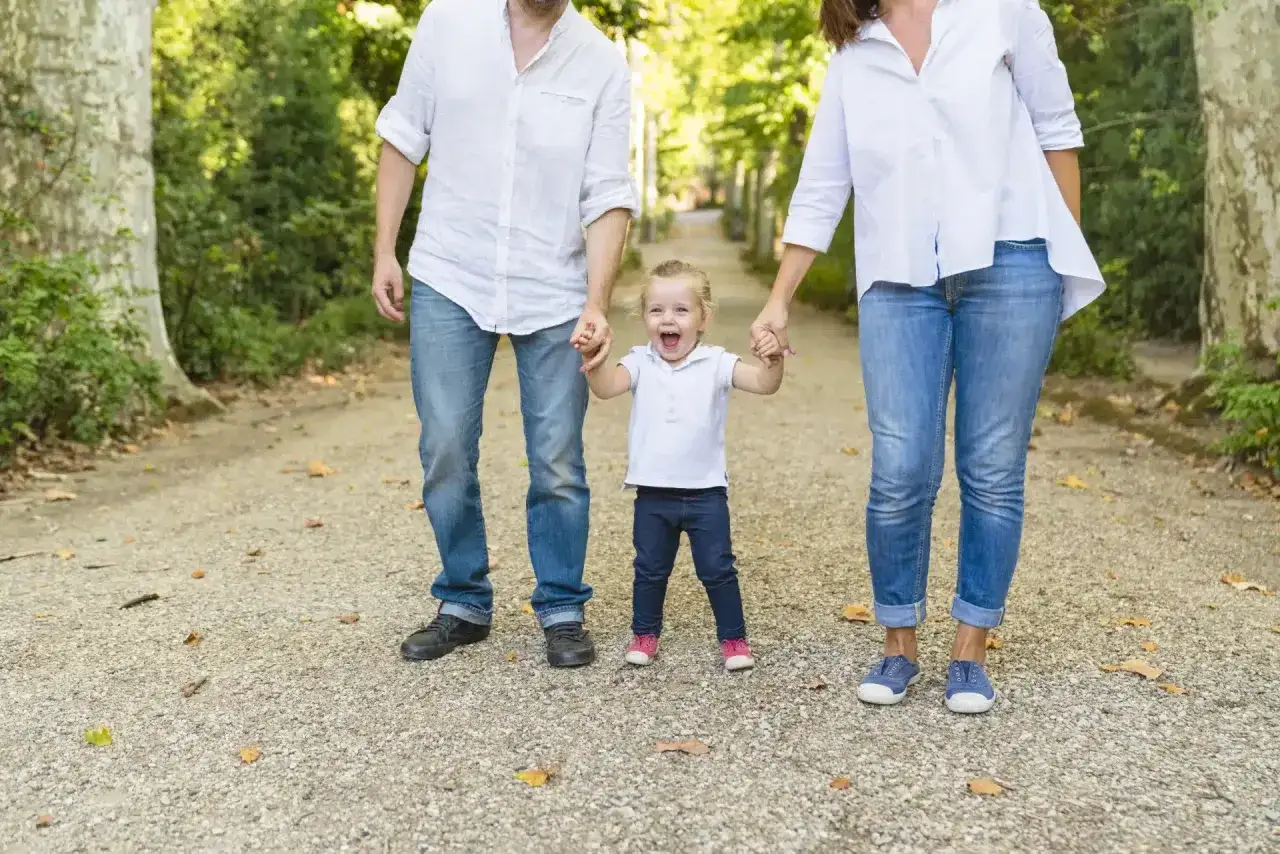 Glückliche Familie mit Kind im Wechselmodell. Vater und Mutter halten Händchen mit ihrem lachenden Kind auf einem Weg im Park.