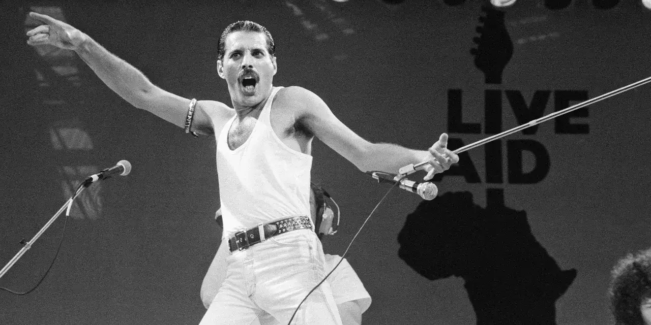 Freddie Mercury na Live Aid, w białej koszulce i spodniach, śpiewa swoje legendarne piosenki.