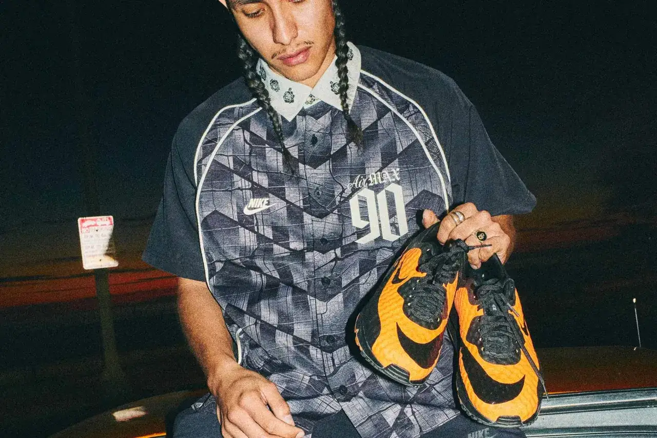 Młody mężczyzna z warkoczami trzyma pomarańczowo-czarne buty Nike Air Max 90. Ma na sobie koszulę z wzorem i logo Nike.