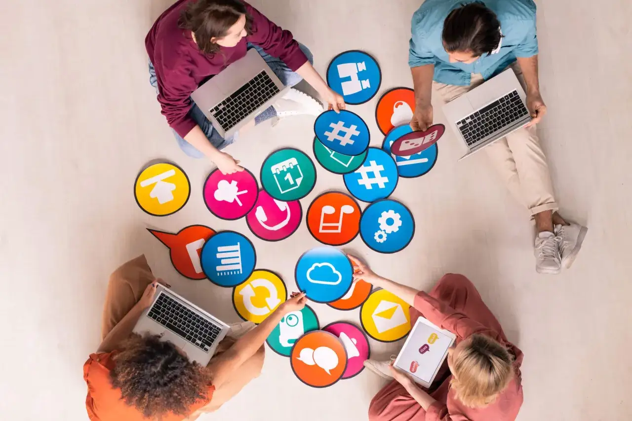 Un grupo de personas colabora en el suelo, rodeado de iconos de aplicaciones de productividad como nube, calendario y música.