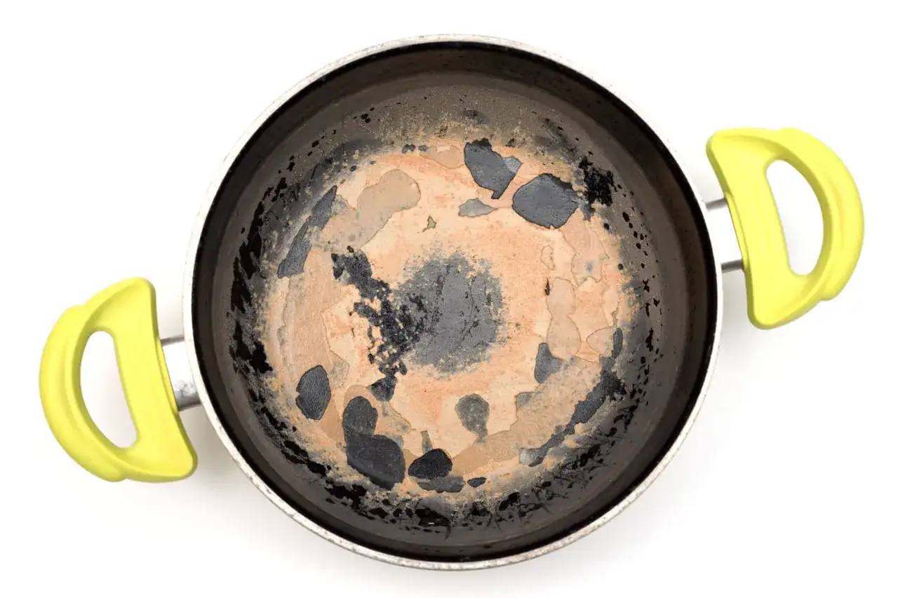 Vue de dessus d'une cocotte minute avec un fond brûlé, marron et noir, taché par la cuisson. Les poignées sont d'un jaune vif.