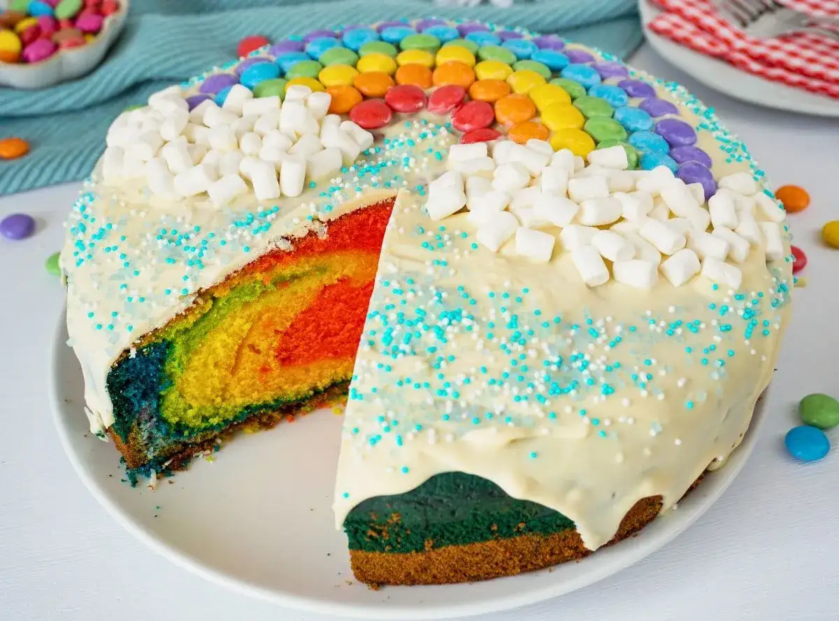 Bunter regenbogen kuchen mit Marshmallows und bunten Streuseln. Ein Stück ist angeschnitten und zeigt die leuchtenden Farben im Inneren.